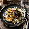 Ein rustikaler Teller mit gegrillten Zitronen-Hähnchen auf einem Reisbett, geröstetem Fenchel und einer Scheibe gegrillter Zitrone, garniert mit frischen Kräutern, auf einem Holztisch mit Silberbesteck und einer braunen Serviette.