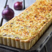 Ein rechteckiger Zwiebelkuchen mit einer goldenen, käsigen Oberfläche aus Hefeteig liegt auf einem Holzbrett. Im Hintergrund sind zwei rote Zwiebeln auf einer hellen Holzfläche zu sehen.