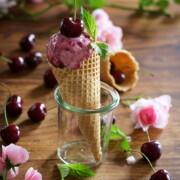 Eine Waffeltüte mit Joghurt-Kirscheis und einer Kirsche obenauf steht in einem Glasgefäß. Frische Kirschen und rosa Blumen sind auf einem Holztisch im Hintergrund verstreut. Perfekt für den Genuss von Eiscreme aus Ihrer Eismaschine!.