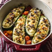 Vier gefüllte Zucchinischiffchen mit einer Käse-Kräuter-Füllung werden mit Kirschtomaten in einer roten Schale gebacken. Dieses rezept gefüllte zucchini vegetarisch sitzt auf einem rotkarierten Tuch auf einem Holztisch, neben einem Holzlöffel.