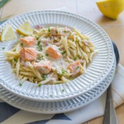 Ein Teller Pasta mit Lachs in cremiger Zitronensauce, garniert mit gehacktem Schnittlauch und Zitronenspalten. Eine Schale mit geriebenem Käse und eine frische Zitrone stehen neben den köstlichen Lachsnudeln auf dem Fliesentisch.