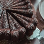 Nahaufnahme eines runden, mit Puderzucker bestäubten Schokoladen-Rotweinkuchens, der auf einem Holzbrett in saubere Stücke geschnitten ist, wobei ein Stück von einem metallenen Kuchenspender angehoben wird.