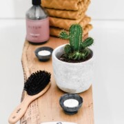 Ein Holztablett hält einen getopften Kaktus, eine Haarbürste, zwei kleine Teelichtkerzen, eine Pumpflasche und gefaltete gelbe Handtücher in einem weiß gefliesten Badezimmer.