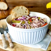 Eine weiße Schüssel mit Nudelsalat, roten Zwiebeln, Paprika und einer Scheibe Brot steht auf einem Holzbrett, das an einen bunten Schweizer Wurstsalat erinnert. In der Nähe stehen Utensilien, zusätzliche Brotscheiben und eine kleine Vase mit gelben Blumen.