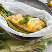 Marinierter Lachs mit grünem Spargel, gebacken in Pergament mit Zitronenscheiben und frischem Dill, serviert in einer Metallschale. Zitronenhälften und Dillzweige liegen auf einem gelben Tuch - ein köstliches Spargelrezept!.