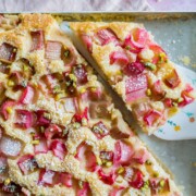 Nahaufnahme eines Blechkuchens mit rosa Rhabarberstückchen und gehackten Pistazien obenauf. Ein Stück dieses köstlichen Rhabarberkuchens wird mit einem blumengeschmückten Spatel aus der Pfanne gehoben.