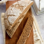 Ein runder Laib rustikales Vinschgauer Fladenbrot, teilweise aufgeschnitten, liegt auf einem hölzernen Schneidebrett mit einem Leinentuch darunter. Eine lange Scheibe liegt neben dem Laib, während im Hintergrund eine Butterdose und ein Messer zu sehen sind.