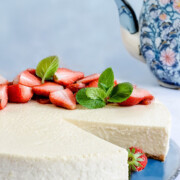 Ein amerikanischer Käsekuchen mit geschnittenen Erdbeeren und Minzblättern liegt auf einem blauen Teller, mit einer geblümten Teekanne im Hintergrund. Ein Stück des cremigen Desserts fehlt.