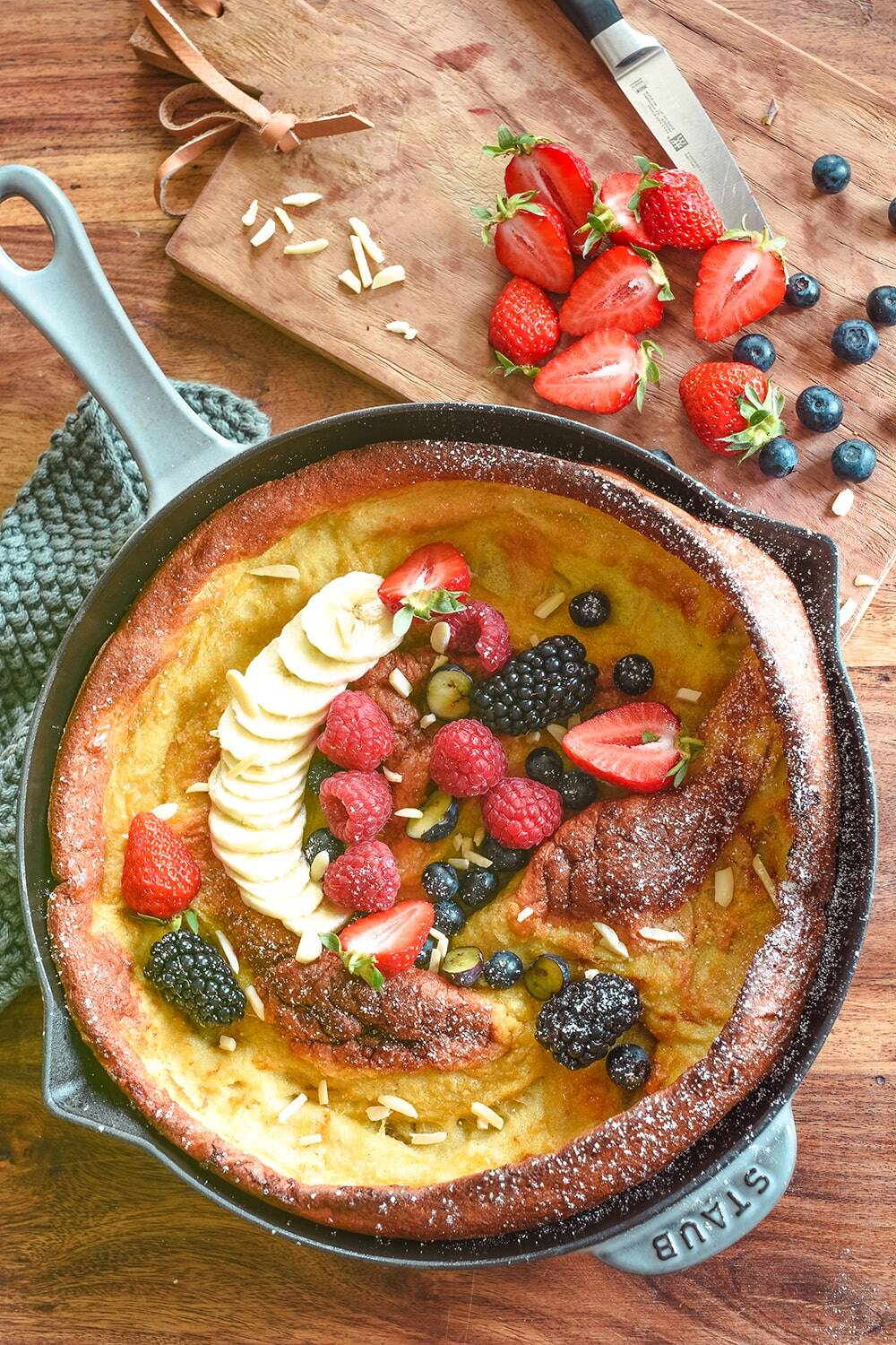 Ein klassischer Dutch-Baby-Pfannkuchen, der in einer gusseisernen Pfanne gebacken und mit in Scheiben geschnittenen Bananen, Erdbeeren, Brombeeren, Himbeeren, Heidelbeeren und Mandelsplittern belegt wird. In der Nähe liegt ein Schneidebrett mit frischen Erdbeeren und einem Messer.