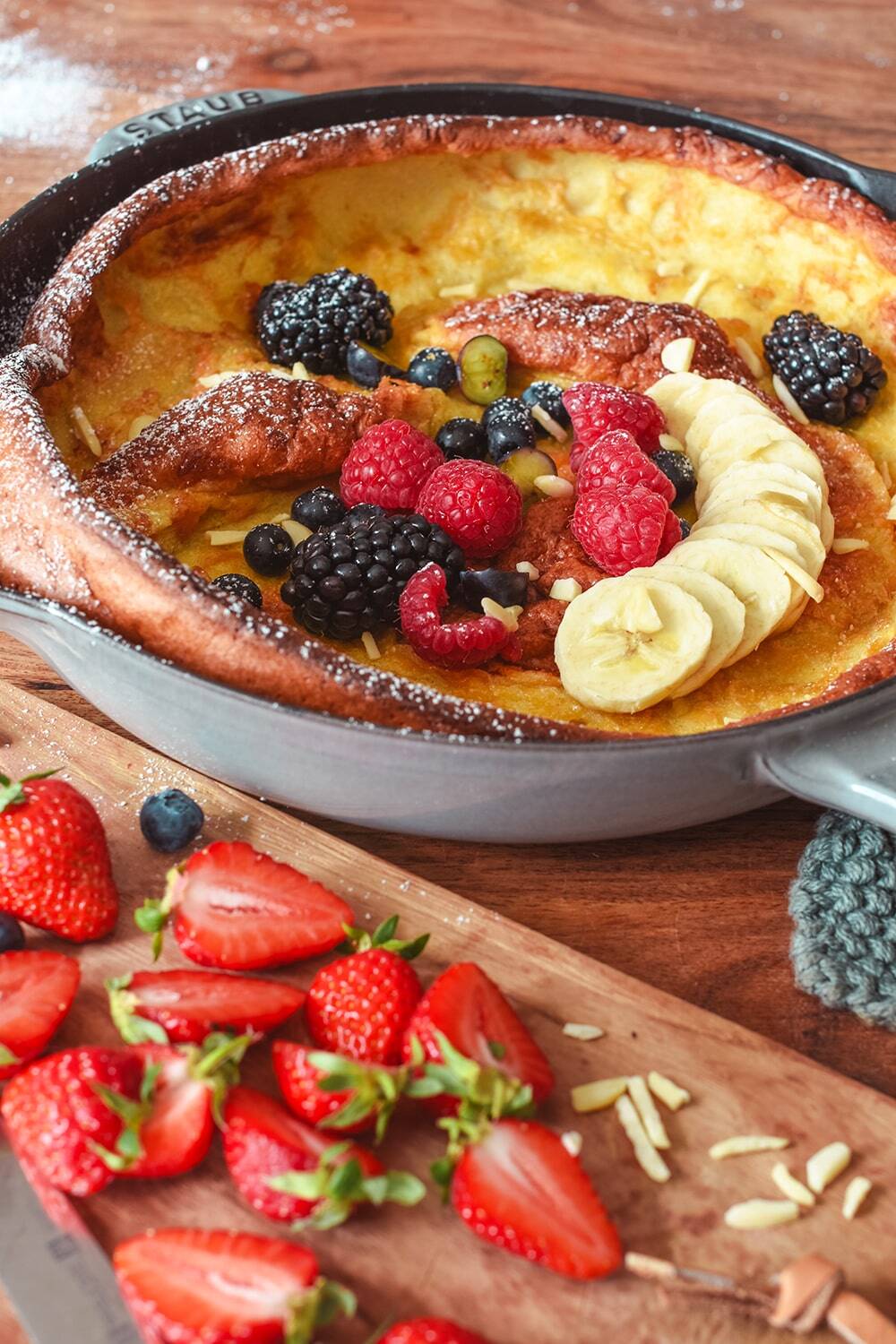 Ein klassischer Dutch-Baby-Pfannkuchen, der in einer Pfanne gebacken und mit Bananenscheiben, Himbeeren, Brombeeren, Blaubeeren und Mandelsplittern belegt wird. Die in Scheiben geschnittenen Erdbeeren liegen auf einem Holzbrett neben dem Dutch Baby.