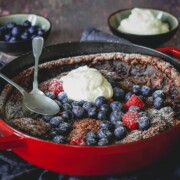 Ein Schoko Dutch Baby Pfannkuchen in einer roten Pfanne, garniert mit Schlagsahne, Blaubeeren und Himbeeren. Zwei Löffel liegen in der Pfanne. Schalen mit zusätzlichen Beeren und Schlagsahne stehen im Hintergrund auf einer dunklen Fläche.