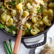 Eine Pfanne mit Fenchel-Forellen-Pasta, geschnittenem Hühnerfleisch, frischen Kräutern und geriebenem Käse, dazu ein Servierlöffel mit Holzgriff. Grüne Zwiebeln und ein Stück Käse liegen daneben auf einer hellen Fläche.