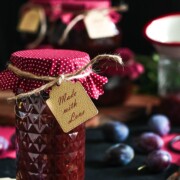 Ein Glas hausgemachter Marmelade mit einem roten gepunkteten Stoffüberzug und einem Etikett mit der Aufschrift Made with Love, umgeben von frischen Zwetschgen und weiteren Gläsern im Hintergrund.