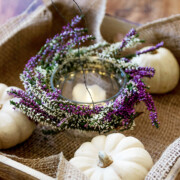 Ein mit lila und weißen Heidekrautblüten verzierter Kerzenhalter aus Glas sitzt in einem Holztablett mit Sackleinen und kleinen weißen Kürbissen. So entsteht eine charmante DIY-Windlichter-Dekoration auf einer Holzfläche.