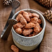 Auf einem braunen Tuch steht eine mit Gebrannten Mandeln gefüllte Keramikschale, in die zwei Metalllöffel eingebettet sind. Im Hintergrund sind Tannenzapfen zur Dekoration angeordnet.