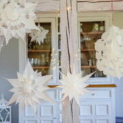 Weiße Papiersternchen, die an Weihnachtssterne und Sterne aus Butterbrottüten erinnern, hängen an einer mit Lichterketten geschmückten Holzleiter, im Hintergrund stehen ein weißer Schrank und Glaswaren.