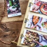 Auf einem Holztisch liegt eine Tafel mit mehreren aneinandergereihten Rezeptkarten, auf denen appetitliche Gerichte abgebildet sind. Eine geflochtene Schnur und eine weitere Rezeptkarte mit einem grünen Getränk sind ebenfalls auf dem Tisch zu sehen.