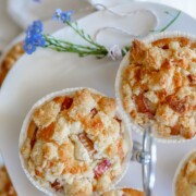 Drei goldene Rhabarber-Streusel-Muffins in weißen Papierförmchen sitzen auf einem weißen Kuchenständer, der mit einem kleinen Zweig blauer Blumen verziert ist. Die Muffins sehen aus wie frisch gebacken und mit Krümeln bedeckt.