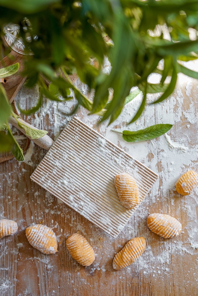 Auf einem mit Mehl bestäubten Holztisch liegen ein geriffeltes Gnocchi-Brett und mehrere ungekochte Süßkartoffel-Gnocchi, die von oben mit grünen Kräuterblättern umgeben sind.