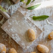 Drei ungekochte Süßkartoffel-Gnocchi liegen auf einem bemehlten Holzbrett. Auf der rustikalen Holzoberfläche sind weitere Gnocchi, Salbeiblätter und Mehl verstreut. Darüber hängen grüne Blätter.