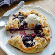 Eine rustikale Blaubeer-Galette, belegt mit gehobelten Mandeln und einer Kugel Vanilleeis, serviert auf einem weißen Teller mit einem Löffel daneben auf einem Holztisch. Eine Scheibe ist herausgeschnitten worden.