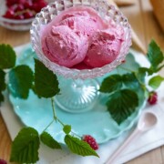 Eine gläserne Dessertschale, gefüllt mit drei Kugeln rosa Johannisbeer-Himbeer-Eis, steht auf einem blauen Teller, umgeben von frischen Himbeeren, grünen Blättern, einer weißen Serviette, einem Löffel und leeren Eiscremetüten auf einem Holztisch.