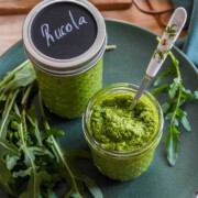 Zwei Gläser mit leuchtendem Rucola-Pesto stehen auf einem grünen Teller, eines offen mit einem Löffel, das andere verschlossen mit einem Deckel mit der Aufschrift Rucola. Frische Raukeblätter liegen neben den Gläsern und verleihen der Szene einen Hauch von Frische.