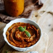 Eine weiße Schale mit cremig-orangefarbenem Auberginenpesto, garniert mit Minzblättern, steht auf einem Holzbrett. Ein Glas mit demselben Auberginenpesto mit allerlei Gemüse und ein braunes Tuch stehen im Hintergrund, daneben ein Löffel.
