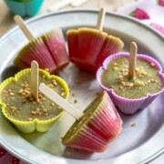 In den bunten Silikonformen stecken Kaktus-Eis-Popsicles, mal grün, mal rosa, jeweils mit einem Holzstäbchen. Diese gesunden Kinder-Eis im Kaktus-Look werden mit zerstoßenen Nüssen bestreut und auf einem runden Metallteller über einem Erdbeertuch serviert.