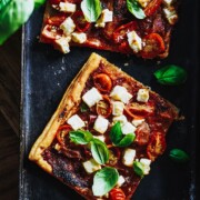 Zwei quadratische Scheiben Blätterteigpizza, belegt mit Kirschtomaten, Fetakäsewürfeln und frischen Basilikumblättern, werden auf einem dunklen Tablett präsentiert, mit zusätzlichen Basilikumblättern in der Nähe, um die Pizza noch lebendiger zu präsentieren.