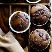 Vier Double Chocolate Muffins mit Schokoladenstückchen in Papiereinlagen liegen auf einem Holztablett, daneben eine braune Stoffserviette und eine Tasse mit Milch.