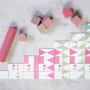 Bunte Holzklötze, die in einem Treppenmuster auf einer hellgrauen Oberfläche angeordnet sind und rosa, grüne und weiße geometrische Muster aufweisen - die perfekte Inspiration für DIY-Dominosteine. Lose Würfel und ein Zylinder sind über dem Arrangement platziert.