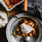 Ein Metallteller mit einer Portion Zwetschgen-Brombeer-Streusel mit Schlagsahne liegt neben einem Löffel und einer Gabel. In der Nähe steht eine Auflaufform mit weiteren Streuseln, und auf dem Holztisch steht eine Schüssel mit Schlagsahne.