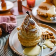 Ein Birnen-im-Schlafrock-Dessert - gebackene Birne in Teig eingewickelt und mit Puderzucker bestäubt - liegt auf einem Teller mit cremiger Soße und Streuseln. Ein weiterer Teller Birne im Schlafrock und eine Gabel sind im Hintergrund auf einem gedeckten Tisch zu sehen.