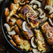 Nahaufnahme einer gusseisernen Pfanne mit gebräunter Maultaschenpfanne, in Scheiben geschnittenen Champignons und in Brühe köchelnden Zwiebeln. Die Gebratenen Maultaschen haben knusprige Ränder und die Pilze sind zart.