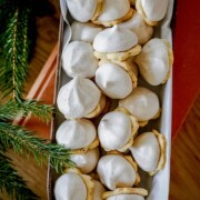 Auf einem rechteckigen, mit Pergamentpapier ausgelegten Tablett liegt ein Stapel Mandelplätzchen mit Zimtbaiserhaube, köstliche Weihnachtsplätzchen mit weißem Baiser und goldenem Boden. In der Ecke sind teilweise immergrüne Zweige zu sehen.