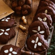 Ein Tablett mit runden Nürnberger Elisenlebkuchen, belegt mit gehobelten Mandeln, steht auf einem Holztisch mit einem Glas Walnüsse, ein paar Erdnüssen und immergrünen Zweigen in der Nähe - eine einladende Variante des klassischen Elisenlebkuchen-Rezepts.