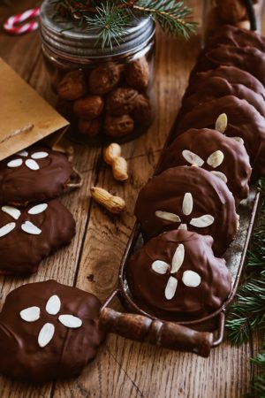 Ein Tablett mit runden Nürnberger Elisenlebkuchen, belegt mit gehobelten Mandeln, steht auf einem Holztisch mit einem Glas Walnüsse, ein paar Erdnüssen und immergrünen Zweigen in der Nähe - eine einladende Variante des klassischen Elisenlebkuchen-Rezepts.
