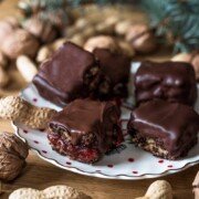 Ein Teller mit fünf schokoladenüberzogenen Schokobrot-Quadraten, von denen einige eine Fruchtfüllung aufweisen, steht umgeben von ganzen Walnüssen und Erdnüssen auf einem Holztisch, mit einem Kiefernzweig im Hintergrund.