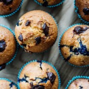Draufsicht auf mehrere goldbraune amerikanische Muffins mit Schokoladenstückchen und Blaubeeren, jeweils in einer blauen Papierhülle, die dicht nebeneinander auf einer Holzfläche angeordnet sind.