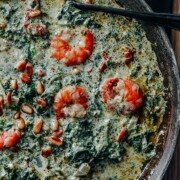 Ein cremiges Spinat-Garnelen-Gericht in der Pfanne, garniert mit Pinienkernen und sonnengetrockneten Tomaten, erinnert an Spinatspaghetti, mit einem Paar schwarzer Stäbchen am Pfannenrand.