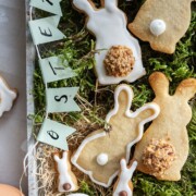 Dekorierte Osterhasen-Plätzchen mit weißem Zuckerguss, kleinen Süßigkeiten und strukturierten Schwänzen ruhen auf grünem Moos neben braunen und weißen Eiern. Ein Papierbanner mit dem Schriftzug OSTERN über den Plätzchen verleiht dieser Osterhasen-Backenszene zusätzlichen Charme.