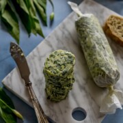 Scheiben von Bärlauchbutter liegen auf einem Marmorschneidebrett neben einem eingewickelten Stück Butter, einem alten Messer, Blattgemüse und Brotscheiben - die perfekte Inspiration für Ihr nächstes Bärlauchrezept auf einer blauen Tischplatte.