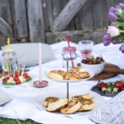 Ein Picknick auf einer weißen Decke mit Blätterteigschnecken und Gebäck auf einem Stufentablett, aufgespießten Snacks, Obst, Kerzen, Getränken und einem Weidenkorb mit lila Tulpen, alles im Freien an einem rustikalen Holzzaun.