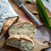 Ein aufgeschnittenes Ciabatta-Brot liegt auf einem Holzbrett, eine Scheibe ist mit Butter bestrichen. Ein Messer liegt daneben, und grüne Blätter sind neben dem Brot verstreut.