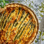 Eine goldbraune Spargelquiche liegt in einer weißen Kuchenform auf einer gemusterten Tischdecke, daneben ein Glas Wasser, ein altes Messer und ein Zweig frischer Kräuter - perfekt für alle, die köstliche Spargelrezepte suchen.