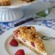 Ein Stück Käsekuchen mit Streuseln und griechischem Joghurt wird auf einem weißen Teller mit zwei frischen Himbeeren und einer Gabel serviert, während der Rest des Kuchens auf einem Ständer im Hintergrund steht.
