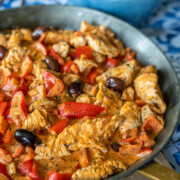 Eine Pfanne mit cremigem Putengeschnetzeltem, rotem Paprika und schwarzen Oliven in einer reichhaltigen Soße. Im Hintergrund steht eine blaue Schale mit gebratenen Kartoffelspalten auf einer blau-weiß gemusterten Tischdecke - ein Vorgeschmack auf griechisches Essen.