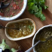 Drei Schalen mit lebhaften Saucen auf einer Holzfläche, daneben frischer Koriander und Auberginen. Perfekt für die Kombination mit Auberginenrezepten oder zum Genießen nach dem Auberginengrillen. Enthält eine rote Salsa, eine grüne Kräutersauce und eine gelbe Sauce auf Ölbasis.