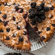 Ein runder Brombeerkuchen, bei dem eine Scheibe fehlt, belegt mit gehobelten Mandeln und frischen Brombeeren, steht auf einem Metalltablett neben einem mit Brombeeren gefüllten Schälchen auf einer weißen Holzfläche - perfekt zum Ausprobieren neuer Brombeerrezepte.